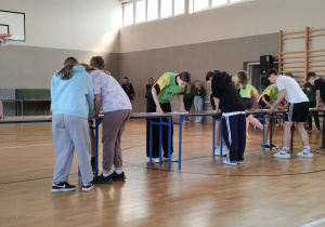 Na sali gimnastycznej stoją stoły. Przy stołach stoją w grupach pochyleni uczniowie. Ubrani są w stroje sportowe.