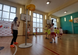 Na sali gimnastycznej widać uczniów ubranych w stroje sportowe - białe koszulki i czerwone spodenki. Po bokach rozstawione są dwa kosze. Uczniowie rozgrywają mecz. Widać piłkę w rękach jednego z dzieci. Widać również po lewej stronie kadru osobę prowadzącą zajęcia.