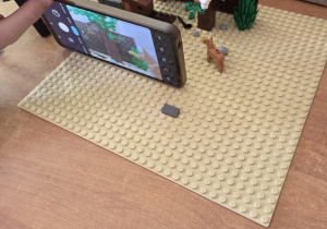 Na stole z klocków zbudowana jest scenka. Widać drzewka, psa, krzaki, ptaszka na gałęzi. Widać również rękę trzymającą telefon z aparatem skierowanym na scenkę z klocków.