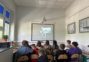 W sali informatycznej dzieci siedza na krzesłach. Na tablicy multimedialnej oglądają lekcję ekologii.