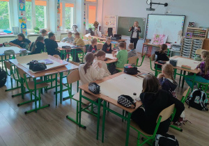 W klasie uczniowie siedza w ławkach. Widać osobę prowadzącą zajęcia. Uczniowie są podzieleni na czteroosobowe grupy.