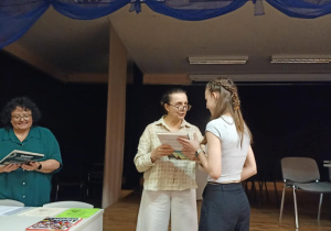 W pomieszczeniu obok stolika organizator wręcza nagrodę naszej uczennicy..