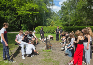 Na trawie w otoczeniu drzew widać uczniów klas ósmych. Siedzą lub stoją z patykami w ręku przy stanowisku z grillem.