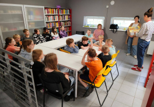 W pomieszczeniu biblioteki przy stołach siedzą uczniowie. Widać osoby prowadzące zajęcia.