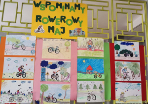 Na kracie na korytarzu wiszą prace uczniów związane z kampanią Rowerowy Maj. Na pracach widać dzieci na rowerach.