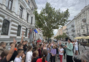 Na ulicy stoją zgromadzone dzieci. Widać osobę trzymającą mikrofon oraz osobę trzymającą aparat.