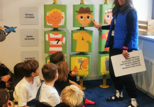 Na niebieskiej podłodze siedzą dzieci. Przed nimi stoi osoba prowadząca zajęcia.
