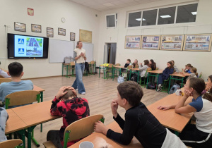 W sali szkolnej siedzą w ławkach uczniowie. Na środku sali widać osobę prowadzącą zajęcia. Z tyłu na tablicy multimedialnej wyświetlona jest prezentacja na temat odblasków.