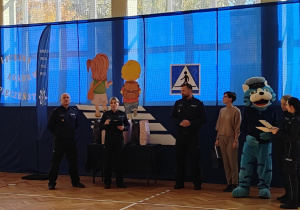 Na sali gimnastycznej stoją policjanci oraz organizatorzy spotkania. Widać maskotkę niebieskiego tygrysa.