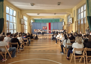 Na sali gimnastycznej siedzą uczniowie ubrani na galowo. W głębi kadru w oddali widać grupę dzieci prowadzącą apel.