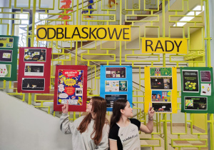 Na kracie na szkolnym korytarzu wiszą prace uczniów wykonane na lekcjach informatyki. Nad pracami wisi napis "Odblaskowe rady". Na środku kadru siedzą dwie dziewczynki i rękami wskazują na plakaty.