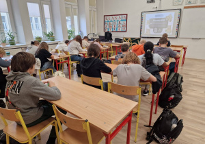 Uczniowie siedzą w ławkach. Rozwiązują zadania z tabliczki mnożenia.