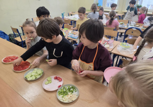 Dzieci siedzą przy ławkach. Na blatach stoją talerze z kolorowymi kanapkami