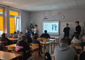 Uczniowie siedzą w klasie w ławkach. W głębi zdjęcia widać osoby prowadzące zajęcia. Na tablicy multimedialnej wyświetlona jest prezentacja.