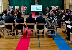 W sali gimnastycznej siedzą uczniowie i goście zgromadzeni na debacie o hejcie. W głębi widać ekran z wyświetlonym plakatem oraz osobę prowadzącą debatę.