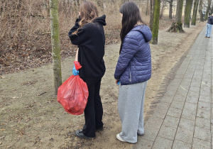 Na alejce w parku stoją dwie dziewczynki. Jedna z nich trzyma różowy worek ze śmieciami.