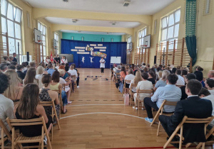 W sali gimnastycznej na krzesłach siedzą uczniowie. W głębi na granatowym suknie wisi dekoracja.