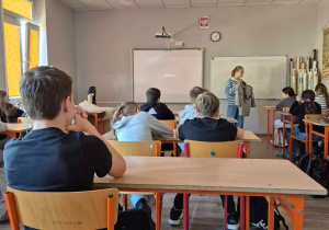 W sali lekcyjnej uczniowie siedzą na krzesłkach. W głębi kadru widać uczennicę prowadzącą zajęcia.