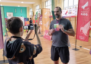 Uczeń robi zdjęcie nauczycielowi wf, który trzyma identyfikator. Nauczyciel jest ubrany w strój sportowy. Za nim znajdują się banery akcji Pho3nix Active School.