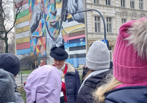 Przed muralem Rubinsteina stoi grupa dzieci. Widać osobę prowadzącą zajęcia.