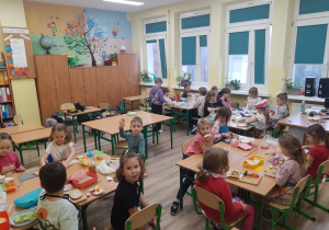 Uczniowie w grupach siedzą przy stolikach. Widać artykuły spożywcze i kanapki leżące na stołach.