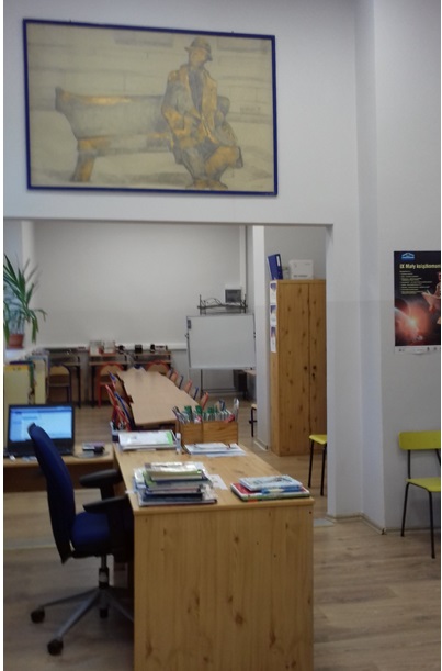na pierwszym planie znajduje się biurko, na ścianie obraz z ławeczką Tuwima, w głębi biblioteki stoły i krzesła
