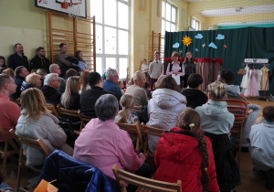 Na sali gimnastycznej siedzą rodzice, uczniowie oraz dzieci. Zgromadzonych wita pani wicedyrektor wraz z dwójką uczniów. W tle widać dekorację do przedstawienia.
