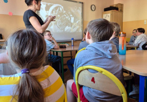 W sali szkolnej uczniowie siedzą na krzesełkach przy stolikach. Po lewej stronie kadru widać osobę prowadzącą zajęcia. W głębi na tablicy multimedialnej wyświetlona jest prezentacja.