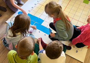 Dookoła maty na podłodze siedzą dzieci. Układają na macie kwadraciki w niebieskim kolorze. Na górze kadru niektóre dzieci trzymają w rękach czerwone i zielone kwadraciki.