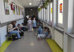 Na szkolnym korytarzu widać uczniów. Po lewej stronie kadru siedzą na ławce. Po prawej stronie kadru siedzą pod oknami na podłodze. Na środku widać idące dwie osoby. Dzieci mają elementy stroju w kolorze niebieskim.