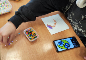 Na zdjęciu widać dziecko siedzące przy stole. Na stole leży telefon z wyświetlonym obrazkiem jajka wielkanocnego. Obok leżą przybory do tworzenia obrazków z koralików.