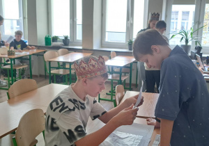 Zdjęcie wykonane w sali lekcyjnej. Z przodu widać ucznia z koroną na głowie siedzącego w ławce. Przed nim stoi drugi uczeń. Z tyłu kadru widać po lewej i po prawej stronie po dwoje uczniów.