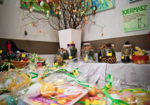 Na stole leżą przedmioty wystawione na sprzedaż na kiermaszu. Są to różne ozdoby wielkanocne. W tle widać gałązki przystrojone kolorowymi jajkami-pisankami. Na ścianie widać wiszące plakaty.