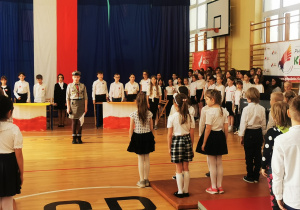 Na sali gimnastycznej w strojach galowych stoją uczniowie. W tle widać powieszoną biało-czerwoną flagę