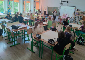 W klasie uczniowie siedza w ławkach. Widać osobę prowadzącą zajęcia. Uczniowie są podzieleni na czteroosobowe grupy.