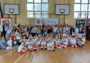 Na sali gimnastycznej stoją uczniowie klas 1-3. Niektórzy ubrani są w stroje sportowe. Za uczniami widać banery akcji "Mały Mistrz"