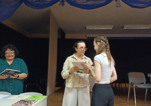 W pomieszczeniu obok stolika organizator wręcza nagrodę naszej uczennicy..