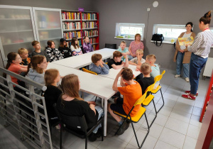 W pomieszczeniu biblioteki przy stołach siedzą uczniowie. Widać osoby prowadzące zajęcia.