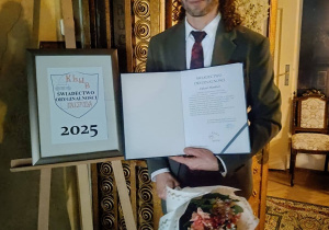 Na zdjęciu p. Handzel trzymający w ręku dyplom i kwiaty, stoi obok sztalugi z plakatem.