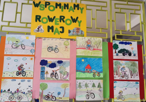 Na kracie na korytarzu wiszą prace uczniów związane z kampanią Rowerowy Maj. Na pracach widać dzieci na rowerach.