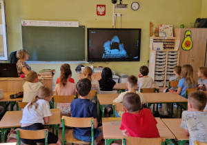 Uczniowie siedzą w klasie. Na telewizorze widać transmisję ze startu rakiety.