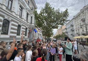 Na ulicy stoją zgromadzone dzieci. Widać osobę trzymającą mikrofon oraz osobę trzymającą aparat.