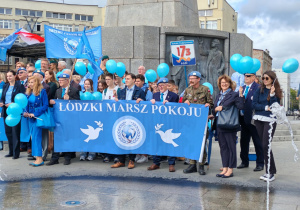 Przed pomnikiem Kościuszki stoją uczestnicy marszu pokoju. Trzymają transparent z gołabkami na niebieskim tle.