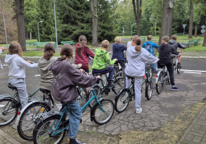 Grupa uczniów siedzi na rowerach. Wszyscy stoją w rzędach na dróżkach motodromu.