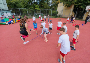 Na boisku szkolnym dzieci ubrane w stroje sportowe skaczą przez skakankę, bawię się chustą