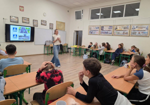 W sali szkolnej siedzą w ławkach uczniowie. Na środku sali widać osobę prowadzącą zajęcia. Z tyłu na tablicy multimedialnej wyświetlona jest prezentacja na temat odblasków.