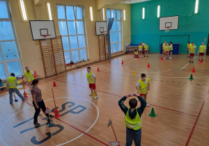 Na sali gimnastycznej widać stojących uczniów. Rozstawione są tory przeszkód. W głębi kadru widać dzieci podzielone na trzy drużyny.
