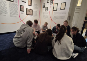 Zdjęcie przedstawia grupę siedmiu młodych ludzi siedzących na podłodze w muzeum. Wokół nich na ścianach znajdują się opisy, teksty i czarno-białe zdjęcia. Grupa zdaje się być zaangażowana w dyskusję lub wspólną pracę, prawdopodobnie w ramach zajęć edukacyjnych.
