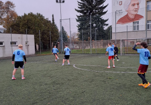 Na boisku stoją zawodnicy ubrani w niebieskie koszulki. Rozgrywają mecz piłki nożnej.