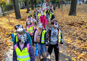 Na ścieżce w parku stoją uczniowie ubrani w kamizelki odblaskowe. Na twarzy mają maski w kształcie wilka.